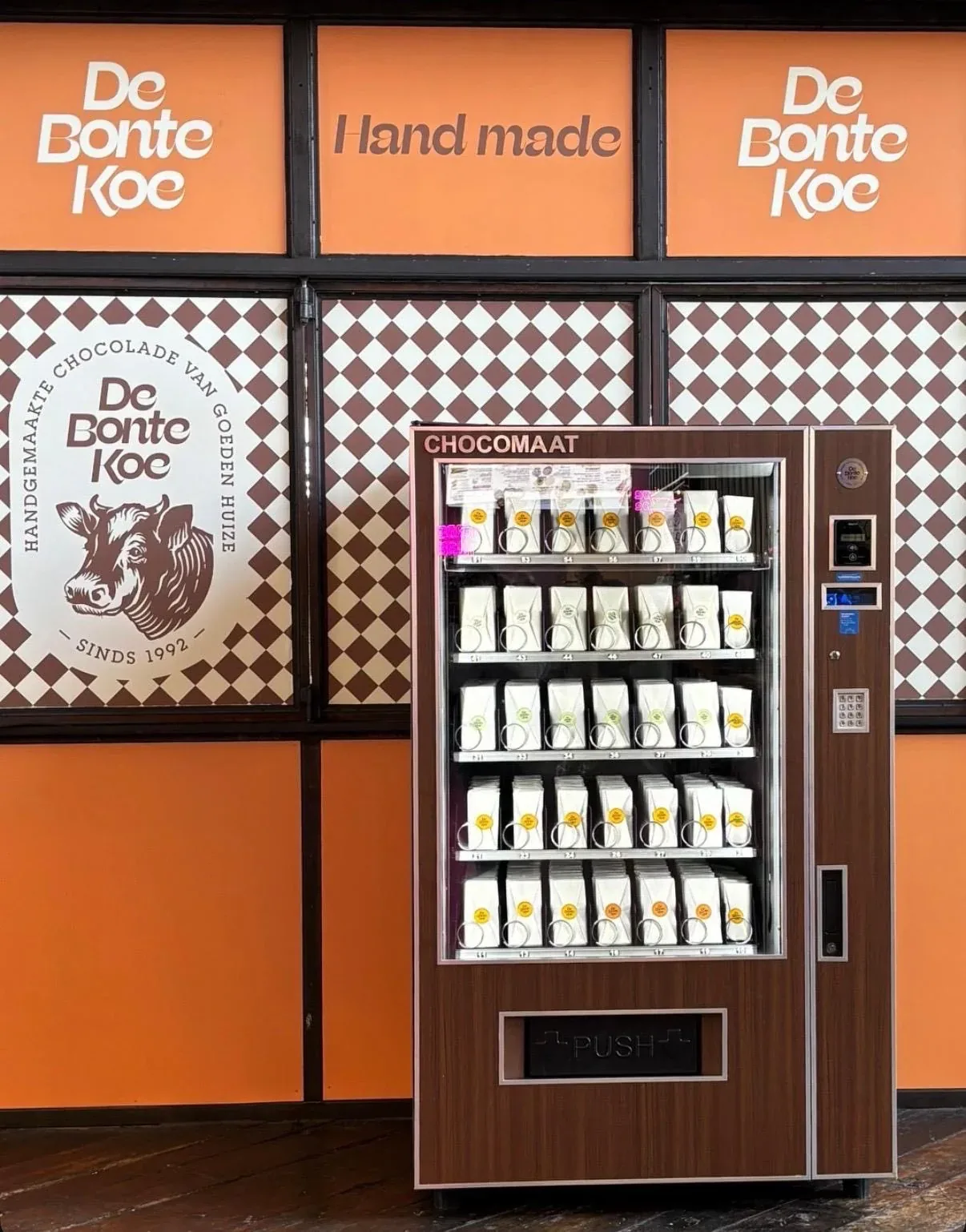 A Chocomaat vending machine full of De Bonte Koe chocolate, against orange and chequered walls.