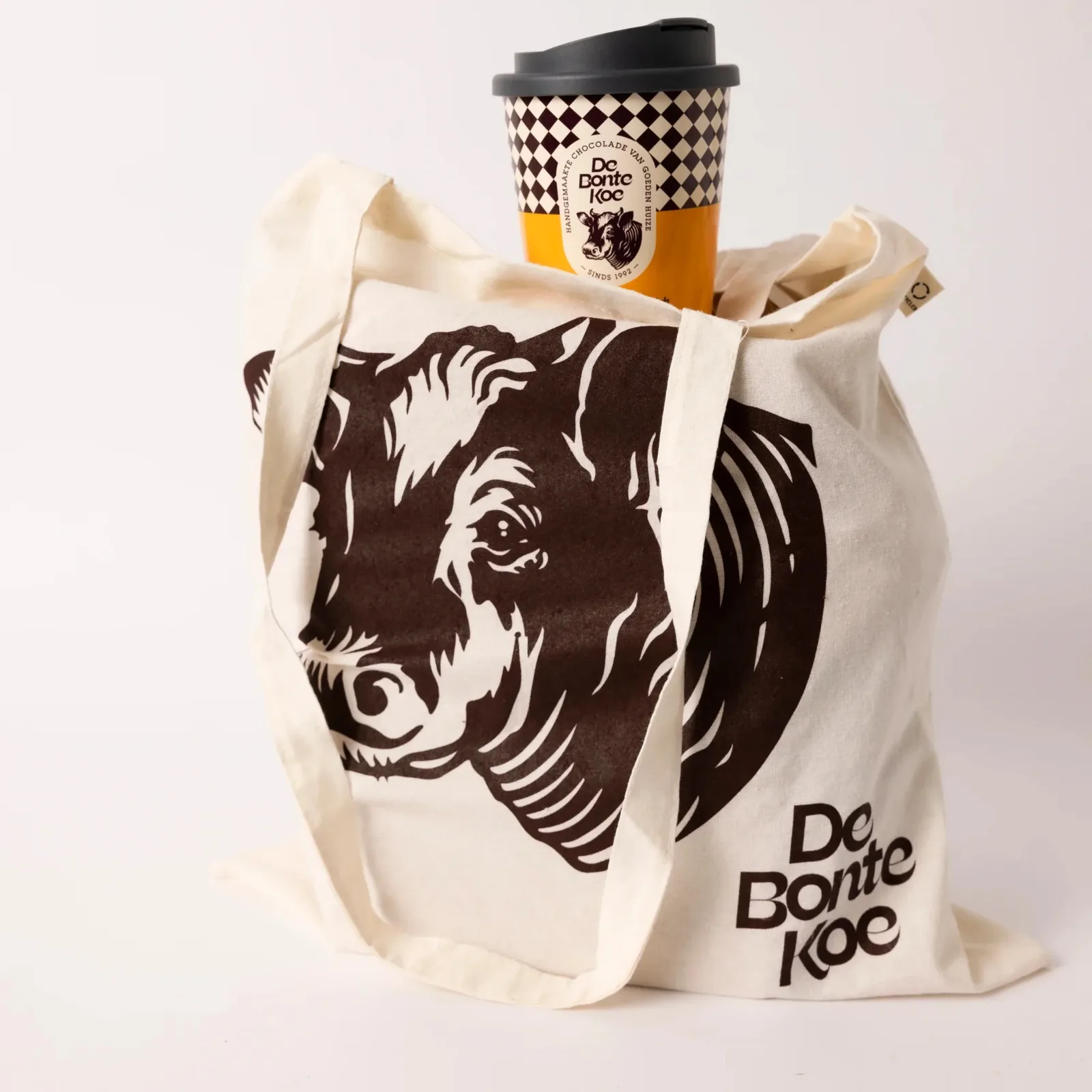 A De Bonte Koe tote bag with the cow logo and a branded coffee cup.